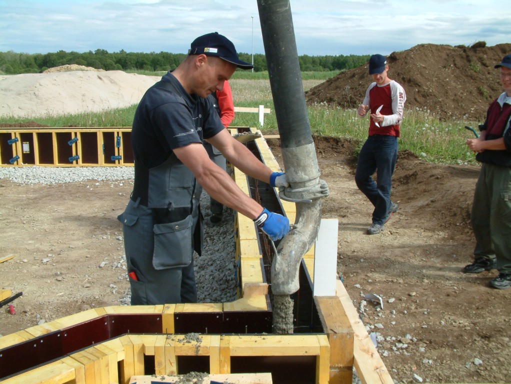 Wylewanie betonu za pomocą pompy do szalunków fundamentowych; pracownik w niebieskich rękawicach kontroluje proces, widoczne zbrojenie i drewniane elementy konstrukcji.
